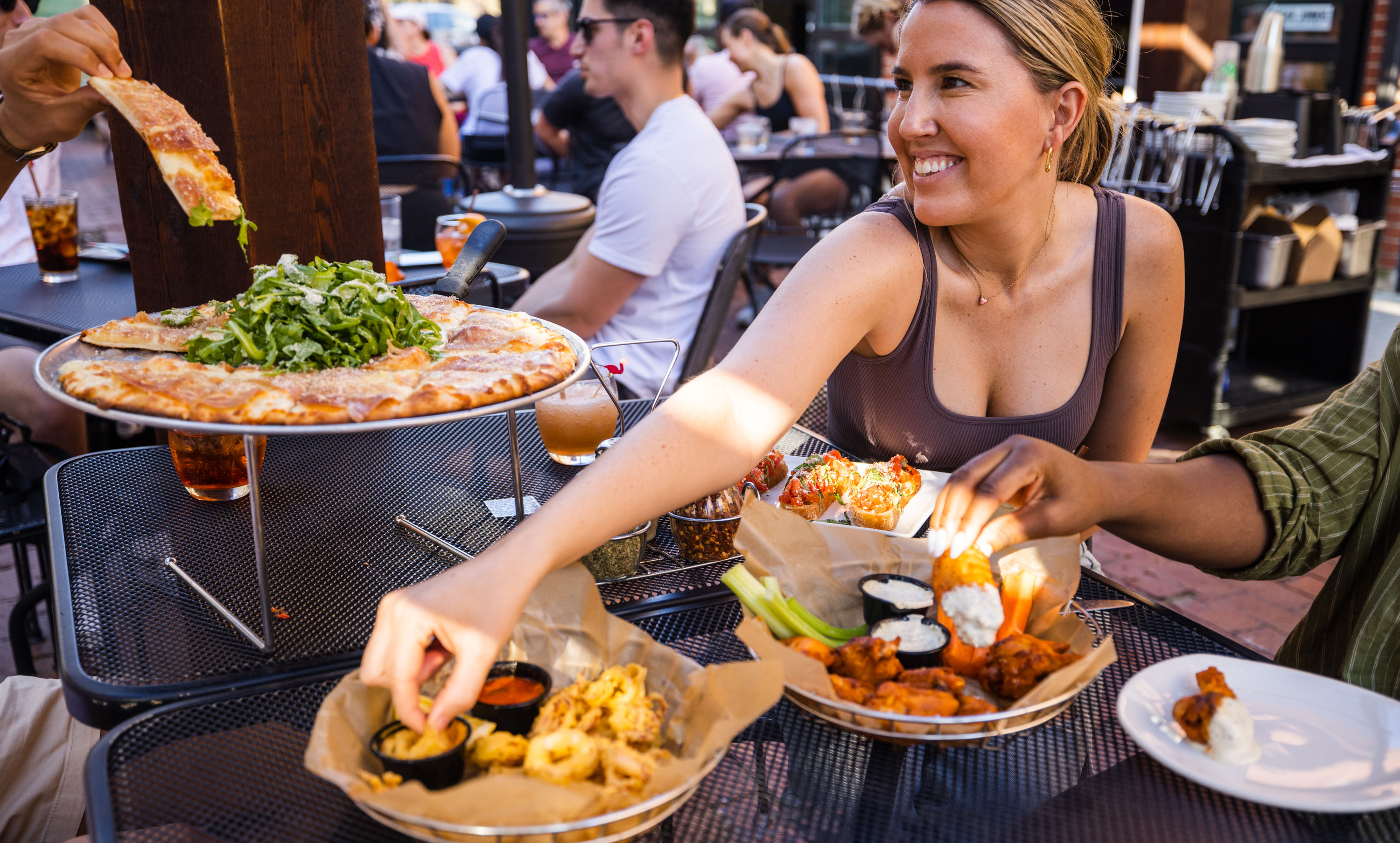 Smiling woman reaches for shared arugula-topped pizza and appetizer plates on an outdoor restaurant patio — wings, fried calamari with dipping sauces and drinks, other diners in the background.