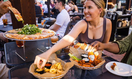 Smiling woman reaches for shared arugula-topped pizza and appetizer plates on an outdoor restaurant patio — wings, fried calamari with dipping sauces and drinks, other diners in the background.