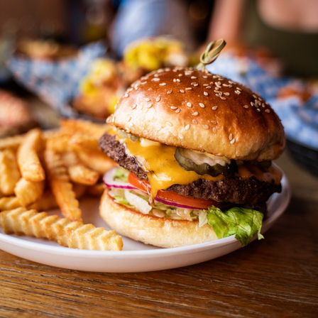 Juicy sesame-seed cheeseburger with melted cheddar, pickles, lettuce, tomato and red onion on a toasted bun, served with crinkle-cut fries on a white plate at a wooden table