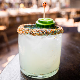 Iced cucumber margarita with zesty chili-salt rim and cucumber wheel garnish on a wooden bar counter, outdoor patio craft cocktail