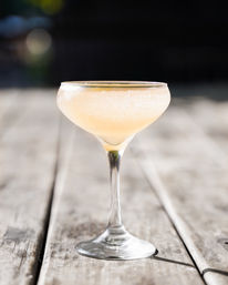Sunlit peach-colored fizzy cocktail in a coupe glass on a weathered wooden patio table with a blurred outdoor background