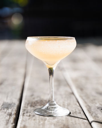 Sunlit peach-colored fizzy cocktail in a coupe glass on a weathered wooden patio table with a blurred outdoor background