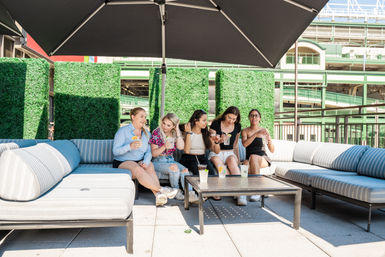 Five friends laughing on an urban rooftop terrace, seated on striped outdoor sofas under a large umbrella, enjoying colorful summer drinks with green hedge panels and city architecture behind them.