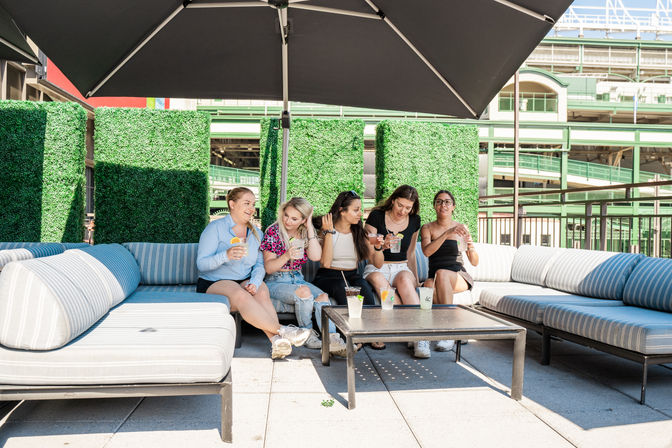 Five friends laughing on an urban rooftop terrace, seated on striped outdoor sofas under a large umbrella, enjoying colorful summer drinks with green hedge panels and city architecture behind them.