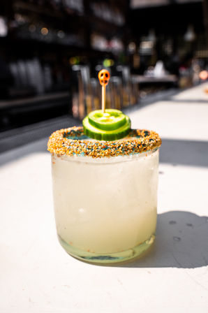 Sunlit cucumber-garnished margarita with crunchy orange chili-salt rim and tiny skull cocktail pick sitting on a bar counter