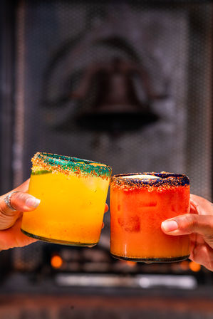 Two vibrant citrus cocktails — one bright yellow, one deep orange — in short glasses with colorful chili-salt rims being clinked by two hands in front of a blurred bar background