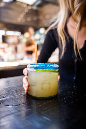 Hand holding a frosty margarita cocktail in a short glass with a bright blue sugar rim and lime slice on a dark wooden bar table, blurred bar interior in background