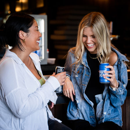 Two friends laughing at a cozy bar counter, casually dressed — one in a denim jacket, the other in a white shirt — each holding a canned beer under warm ambient lighting.