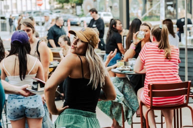 Playful woman in a tan baseball cap striking a pose on a busy outdoor patio brunch in the city, friends chatting and sipping drinks at nearby tables.