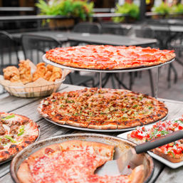 Al fresco pizzeria spread on a wooden patio table: stacked thin-crust pepperoni and specialty pizzas, a basket of garlic knots and tomato bruschetta.