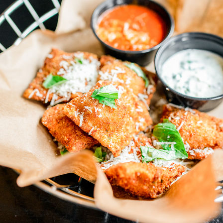Crispy breaded fried ravioli sprinkled with grated Parmesan and basil in a parchment-lined basket, served with marinara and herbed ranch dipping sauces — Italian appetizer.