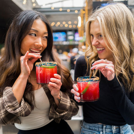Two friends laughing and sipping bright red cocktails with colorful sugar rims and lime wedges at an urban patio bar with string lights