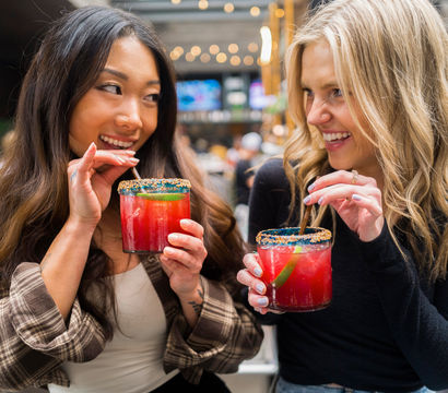 Two friends laughing and sipping bright red cocktails with colorful sugar rims and lime wedges at an urban patio bar with string lights