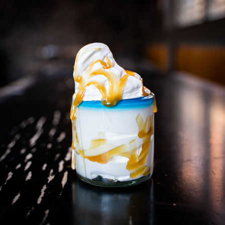 Close-up of caramel-drizzled soft-serve dessert in a small glass jar with a blue rim, whipped cream swirls overflowing onto a dark wooden table in a blurred cafe setting.