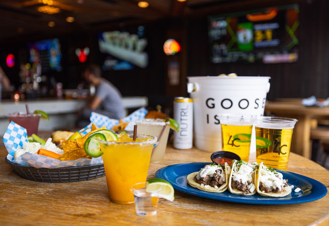 Three street tacos with beef and crema on a blue plate, basket of chips and dip, bright mango margarita with lime and a shot glass, and two beers on a wooden table in a lively sports bar with TV screens