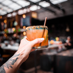 Tattooed hand holding a spiced orange craft cocktail with a colorful crunchy rim and straw in a lively bar interior.