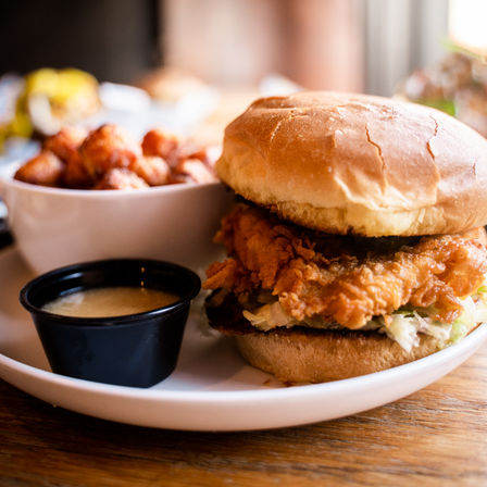 Mouthwatering crispy fried chicken sandwich on a toasted bun with shredded lettuce, served with dipping sauce and tater tots on a white plate on a wooden table in a casual restaurant