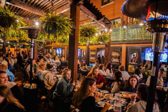 Bustling urban outdoor dining patio at night — wooden pergola with hanging fern planters, string lights and patio heaters, groups enjoying food beside a brick building
