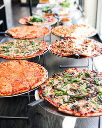 Mouthwatering assortment of thin-crust pizzas — plain cheese, meat, and veggie with green peppers and mushrooms — displayed on metal stands along a pizzeria counter.