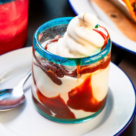 Close-up of a caramel- and chocolate-drizzled vanilla soft-serve dessert in a short glass with a turquoise rim, served on a white plate with a spoon.