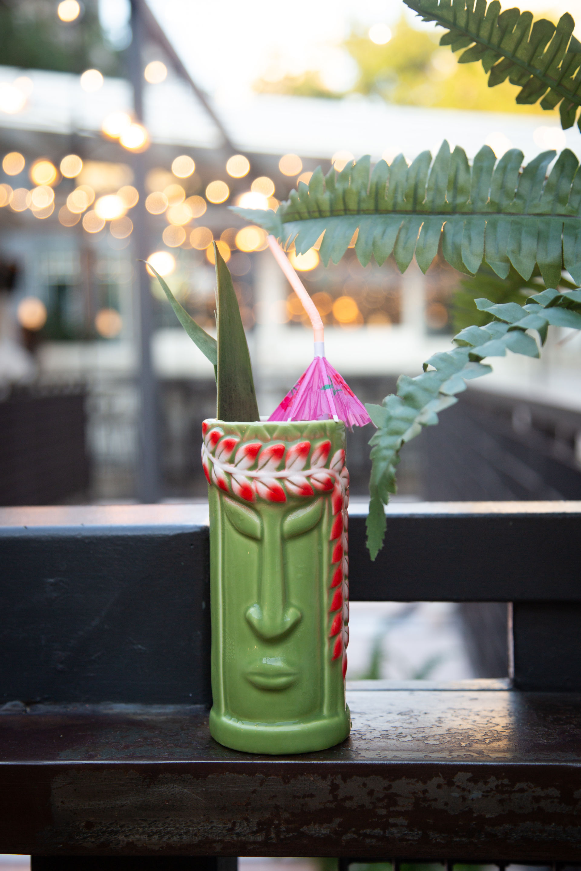 Green ceramic tiki mug with red leaf garland, pink cocktail umbrella and straw on an outdoor patio railing, fern leaves nearby and warm bokeh string lights in the evening background