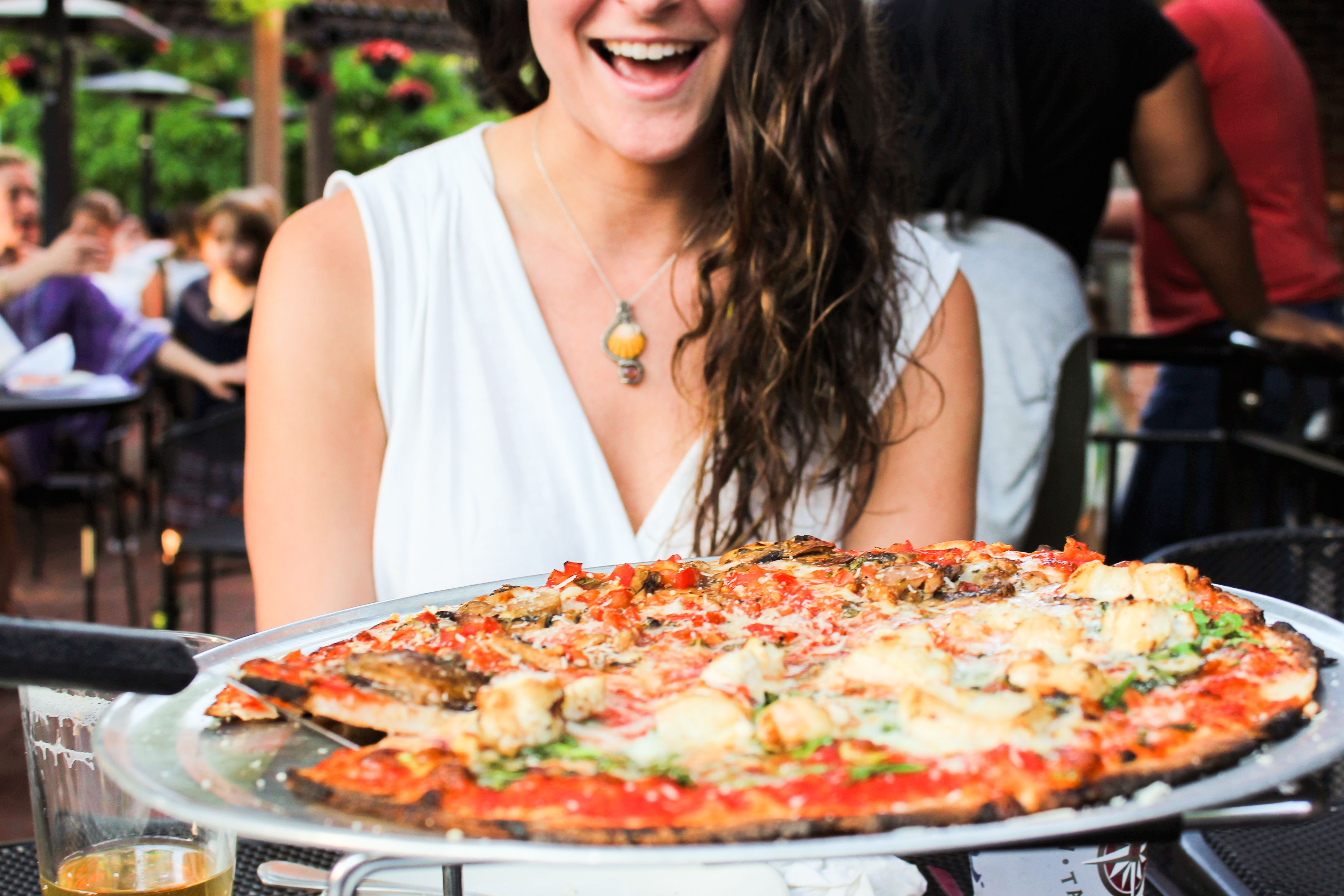 Smiling woman in a white top delighted by a large thin-crust pizza with melted cheese and fresh toppings served on a metal tray at an outdoor restaurant patio