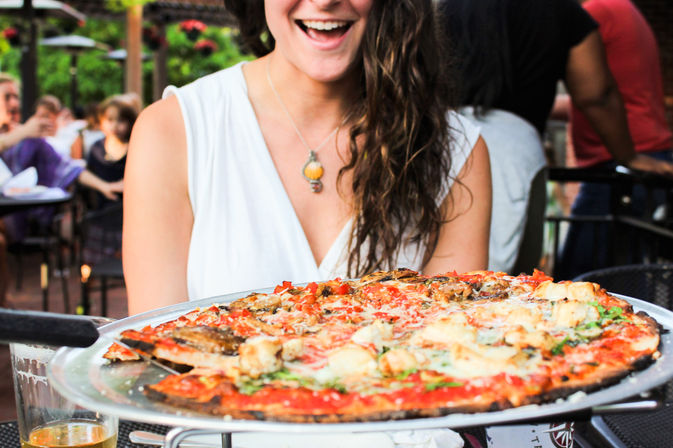 Smiling woman in a white top delighted by a large thin-crust pizza with melted cheese and fresh toppings served on a metal tray at an outdoor restaurant patio