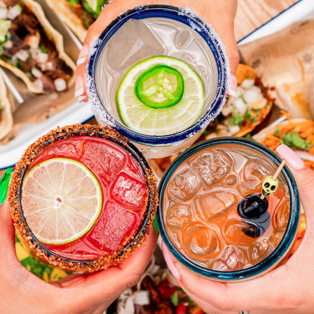 Top-down view of three festive cocktails held over a tray of tacos — a lime-and-jalapeño margarita with a salted rim, a chili-rimmed red cocktail with a lime slice, and an iced amber cocktail garnished with an olive.