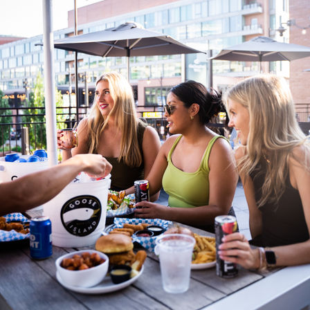 Three friends laughing on a downtown rooftop patio under umbrellas, sharing burgers, fries and canned drinks from an ice bucket during a sunny summer meal.