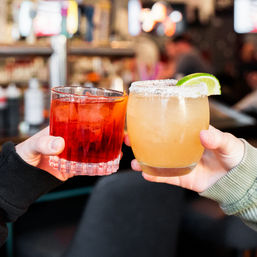 Two craft cocktails clinking at a busy bar — a deep red drink in a rocks glass and a salt-rimmed citrus cocktail with a lime wedge in a short glass, blurred bar background.