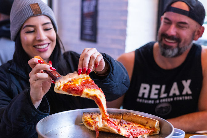 Two friends sharing a gooey Chicago-style deep-dish pepperoni pizza in a casual pizzeria; a woman lifts a cheesy slice from a metal pan.