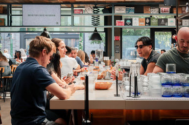 Bustling downtown industrial-style café bar with communal counter seating, patrons chatting and scrolling phones, stacked glassware, bowls and drinks under pendant lights and large street-facing windows.