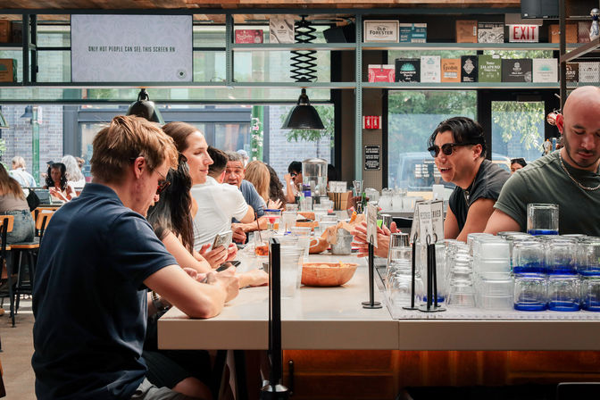 Bustling downtown industrial-style café bar with communal counter seating, patrons chatting and scrolling phones, stacked glassware, bowls and drinks under pendant lights and large street-facing windows.