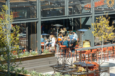 Sunny urban al fresco patio with groups dining at orange metal tables and chairs under string lights and patio heaters, industrial metal framework, wooden barrels and succulent planters.