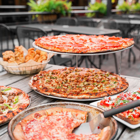 Mouthwatering outdoor patio spread of large thin-crust pizzas on a two-tier stand—pepperoni and veggie varieties—with a pan of sliced pizza, a basket of fried chicken, and tomato bruschetta on a wooden table.