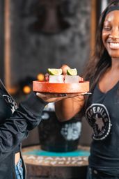 Bar scene: two large ice-shot cubes topped with lime wedges on an orange tray being passed between hands, smiling server and blurred bar background.