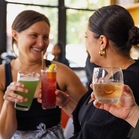 Smiling friends toasting colorful cocktails and a glass of white wine in a sunlit urban cafe bar with large windows.
