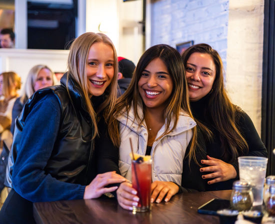 Three smiling women leaning at a bar counter with a red, fruit-garnished cocktail — casual night out at a cozy indoor restaurant.