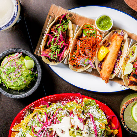 Overhead shot of colorful Mexican tacos and loaded nachos: three soft tacos (fish, shredded meat, veggie) on an oval plate with green salsa, a bowl of fresh guacamole with lime and radish, loaded nachos with cheese and pickled onions, and a margarita with lime and chili-rim.