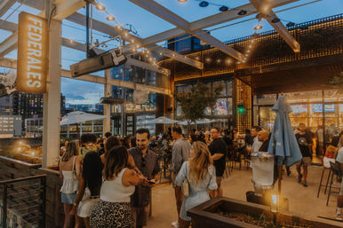 Busy downtown rooftop bar patio at dusk with string lights, umbrellas, high-top tables and groups socializing against a city skyline backdrop.