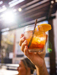 Hand holding an orange-garnished iced cocktail in a short glass with straw, raised against a sunlit outdoor patio with lens flare