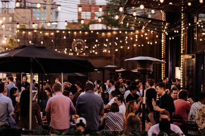 Lively urban outdoor patio bar at dusk with warm string lights, umbrellas and patio heaters, groups of people socializing and enjoying drinks