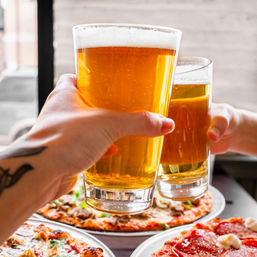 Two hands toasting pints of golden craft beer over pepperoni and veggie pizzas at a casual pizzeria