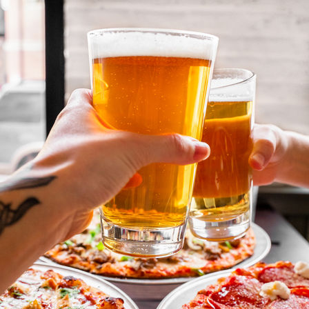 Two hands toasting pints of golden craft beer over pepperoni and veggie pizzas at a casual pizzeria
