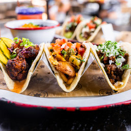 Taco trio close-up: three colorful Mexican street tacos in a metal rack — fried chicken with pickles, grilled fish with corn salsa and pico, and seasoned beef topped with onion and cilantro.