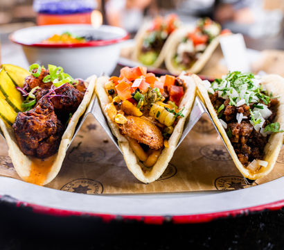 Taco trio close-up: three colorful Mexican street tacos in a metal rack — fried chicken with pickles, grilled fish with corn salsa and pico, and seasoned beef topped with onion and cilantro.
