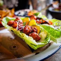 Crispy fried chicken lettuce wraps with pickled cucumber slices and shredded purple cabbage, served as a colorful appetizer on a restaurant plate.