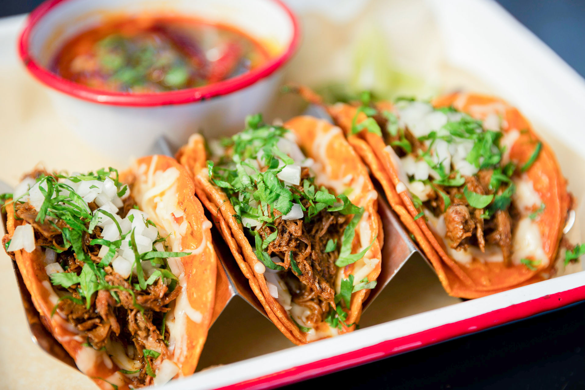 Three Mexican braised beef street tacos in toasted corn tortillas topped with chopped onion, cilantro and melted cheese, served with a side bowl of red salsa