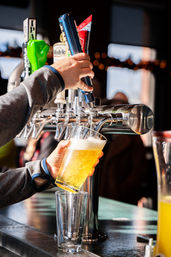 Hand pulling a beer tap handle pouring a frothy golden draft pint into a tilted glass at a lively bar counter
