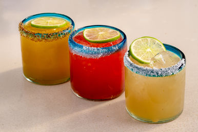 Vibrant trio of margarita-style cocktails in blue-rimmed short glasses with salted rims and lime slices — red and yellow drinks on a light surface.